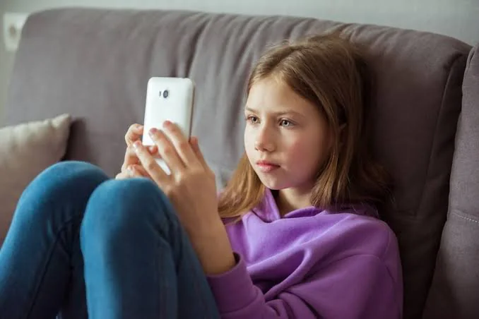 A pre-teen girl in a purple hoodie sitting on a sofa absorbed in her smartphone, illustrating a child's heavy screen-time and phone use at home