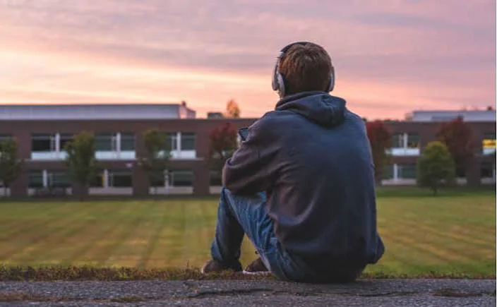 A teenage boy in a hoodie and headphones sitting alone on a curb outside a school building at dusk, gazing across an empty field, conveying loneliness and having no friends