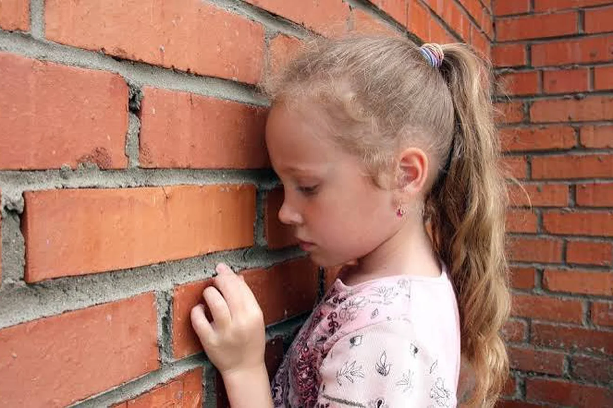 A young girl with a ponytail leaning her forehead against a red brick wall, looking down with a quiet, discouraged expression