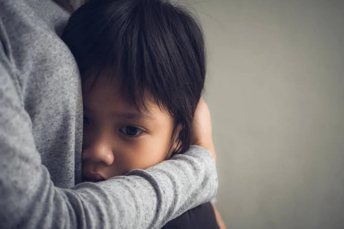 A young child with dark hair resting their head on a parent's shoulder, clinging closely and looking anxious and withdrawn